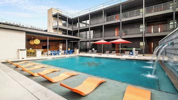 A modern pool area with lounge chairs and umbrellas, a bar area, and a multi-story building with balconies.