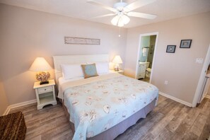 A bedroom with a bed, two bedside tables, a ceiling fan, a wall-mounted light, and a door leading to a bathroom. - Amelia Island Ocean View + Pool Top Floor (Fernandina Beach)