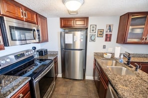 A kitchen with stainless steel appliances, granite countertops, wooden cabinets, and a tiled floor. - Drift Away Beachfront 2BR w/ Balcony & Pool (Fernandina Beach)