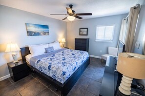 A bedroom with a bed, nightstands, a ceiling fan, a dresser, a window with blinds, and a wall-mounted picture. - Drift Away Beachfront 2BR w/ Balcony & Pool (Fernandina Beach)