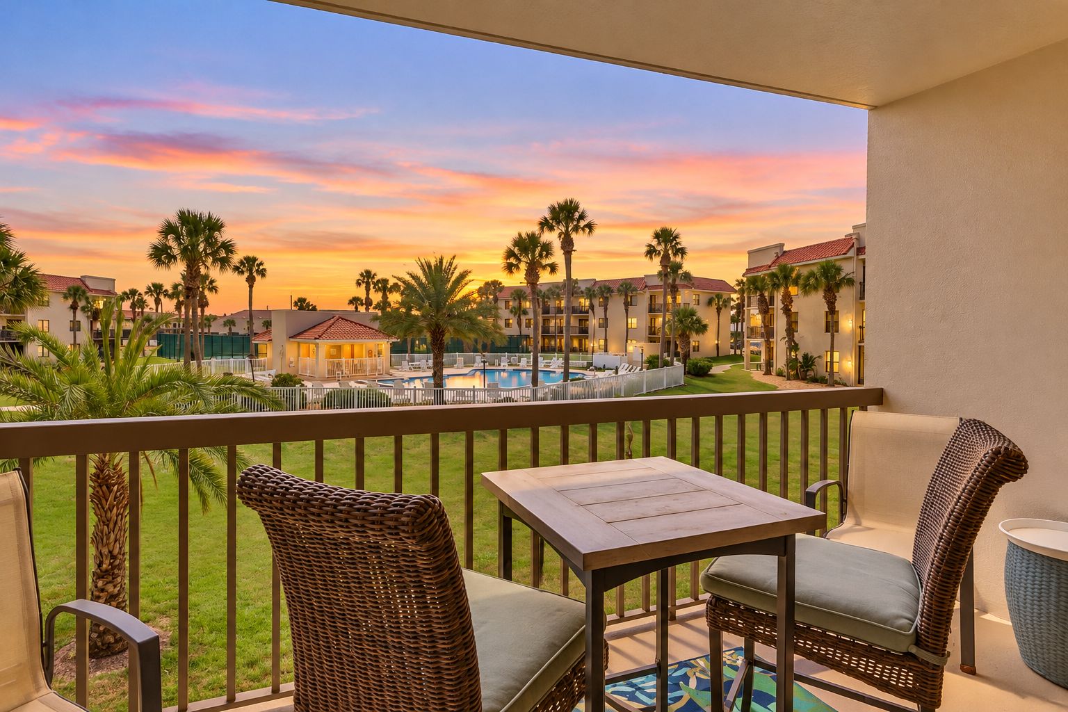 A balcony with wicker furniture, a pool, and a view of a resort with palm trees and buildings.