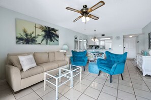 A living room with a beige sofa, two blue armchairs, a glass coffee table, a ceiling fan, and a kitchen in the background. - Coastal Condo Pool View + Heated Pool (St. Augustine)