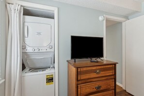 A laundry room with a washing machine and dryer, a television on a wooden dresser, and a door leading to another room. - Beachfront 2BR Pool Direct Access (Fernandina Beach)