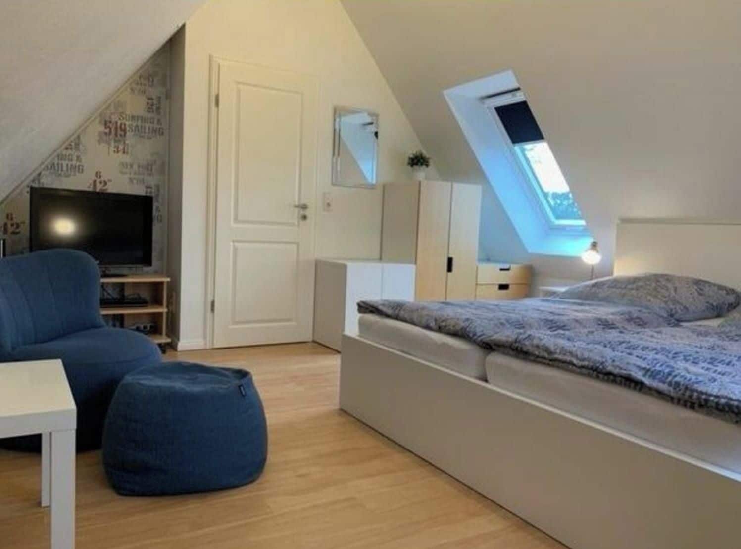 Quarto compacto com cama, televisão, escrivaninha e um pequeno espaço para sentar.