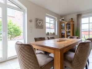 A dining area with a wooden table and cushioned chairs, a heart-shaped artwork on the wall, and a view of the outside. - Holiday Home With 2 Bedrooms (St. Peter-Ording)