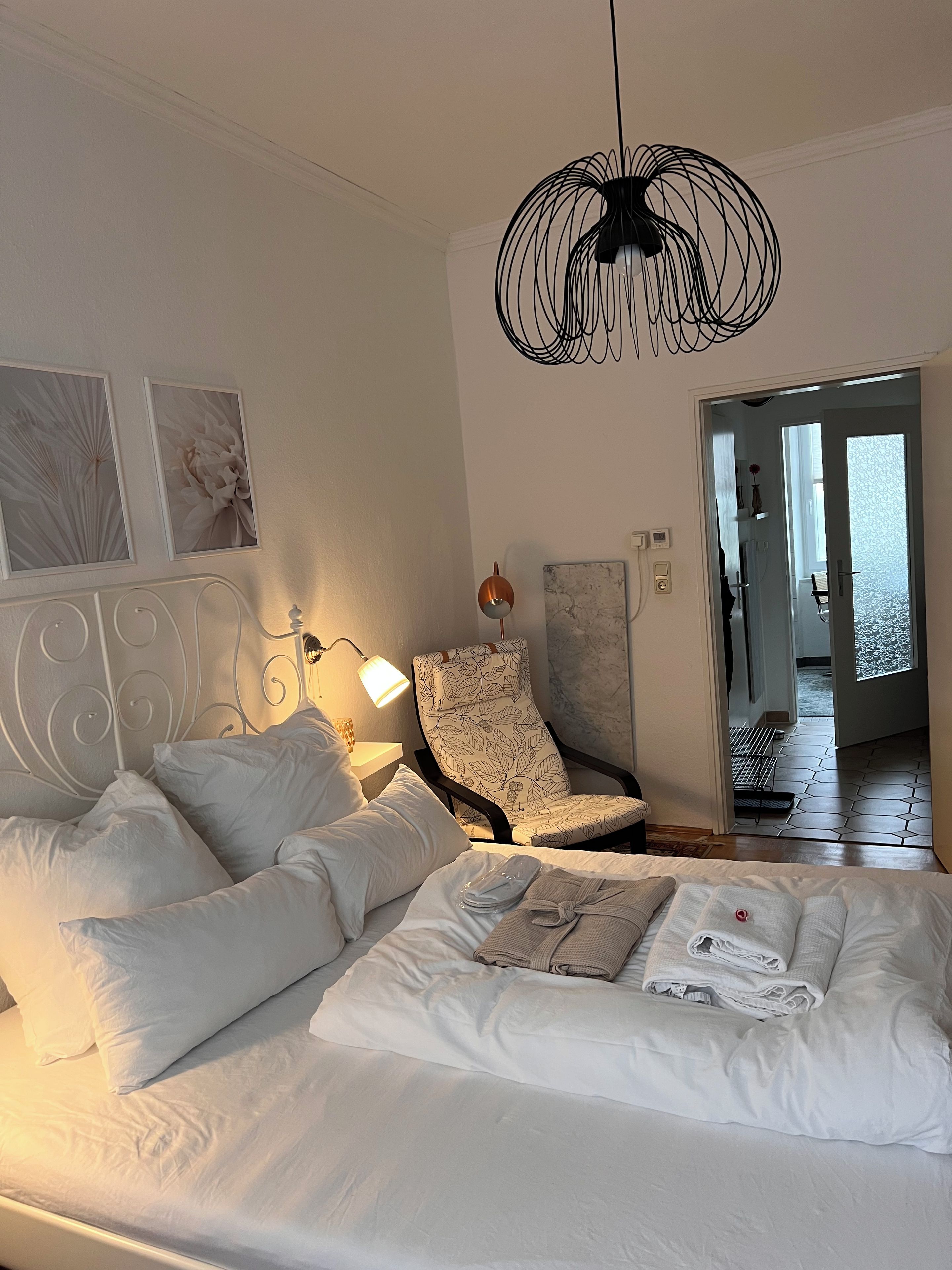 Ein ordentlich bezogenes Bett mit Kissen und einem gefalteten Handtuch, ein Stuhl mit Blumenmuster, eine Wandleuchte und eine einzigartige schwarze Pendelleuchte.