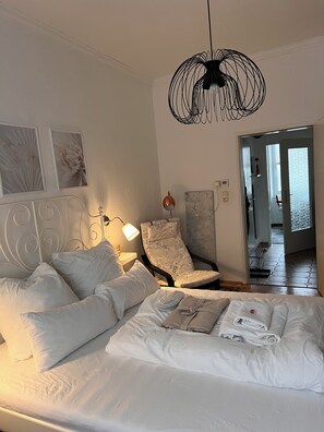 A neatly made bed with pillows and a folded towel, a chair with a floral pattern, a wall-mounted lamp, and a unique black pendant light. - Romantic vacation rental with bathrobes—right in the heart of downtown (Bamberg)