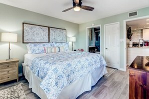A bedroom with a bed, bedside tables, a ceiling fan, and a view into the adjacent kitchen. - Ocean View 2BR Pool Walk to Beach (Fernandina Beach)
