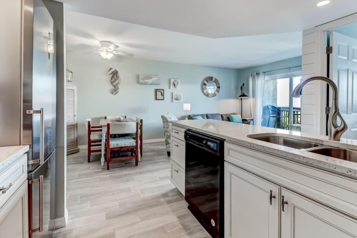 Cocina moderna con lavavajillas, comedor con mesa y sillas de madera, y vista al exterior por una ventana.