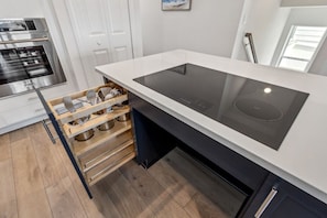 A modern kitchen with a built-in induction cooktop, wooden storage shelves, and stainless steel appliances. - Brand New 4BR Ocean Views Pool (Fernandina Beach)