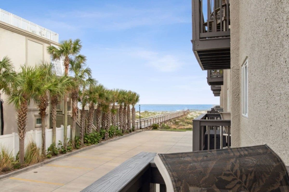 A pathway lined with palm trees leading to a beach, with a building on the left and a balcony on the right.