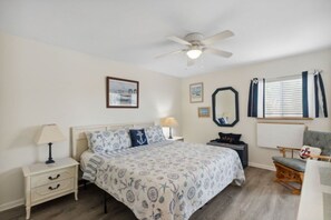 Een slaapkamer met een bed, nachtkastje, lamp, stoel, plafondventilator en kunstwerken aan de muren.
