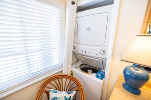 A compact laundry room with a washing machine and dryer, a wicker chair, and a lamp. - Closest to Beach in Sanddollar! Beachfront w/ Pool (Fernandina Beach)