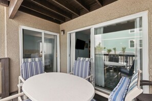 A covered patio with a round table and two striped chairs, a sliding glass door, and a view of a building across the street. - Beachfront Condo Pool Sleeps 6 (Fernandina Beach)