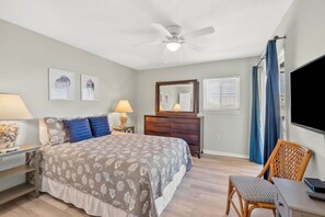 Ein Schlafzimmer mit einem Bett, einer Kommode, einem Sessel, einem Schreibtisch, einem Deckenventilator und einem Fernseher.