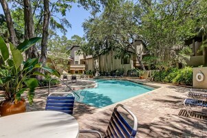 A pool area with lounge chairs and a table, surrounded by trees and buildings.