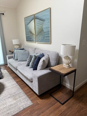 A modern living room with a grey sofa, multiple cushions, a small side table with a lamp, and a large painting above the sofa. - Dog-friendly 3BR Walk to Beach Pool (Fernandina Beach)