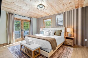 Ein Schlafzimmer mit Holzbalkendecke, einem großen Bett mit weißer Bettwäsche, einer Bank mit Handtüchern, einem Nachttisch mit Lampe und einem Fenster mit Vorhängen.