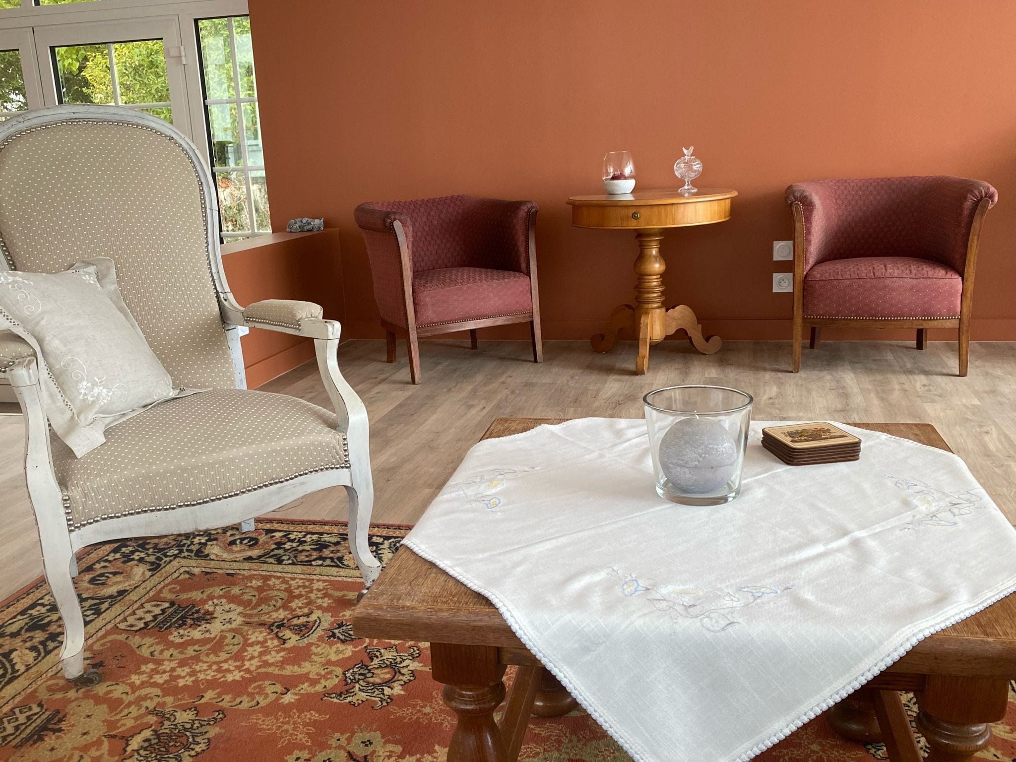 Una habitación con un alfombra estampado, una silla blanca, dos sillones tapizados en rojo, una mesa de madera con un vaso y un libro, y una mesita redonda con dos copas.