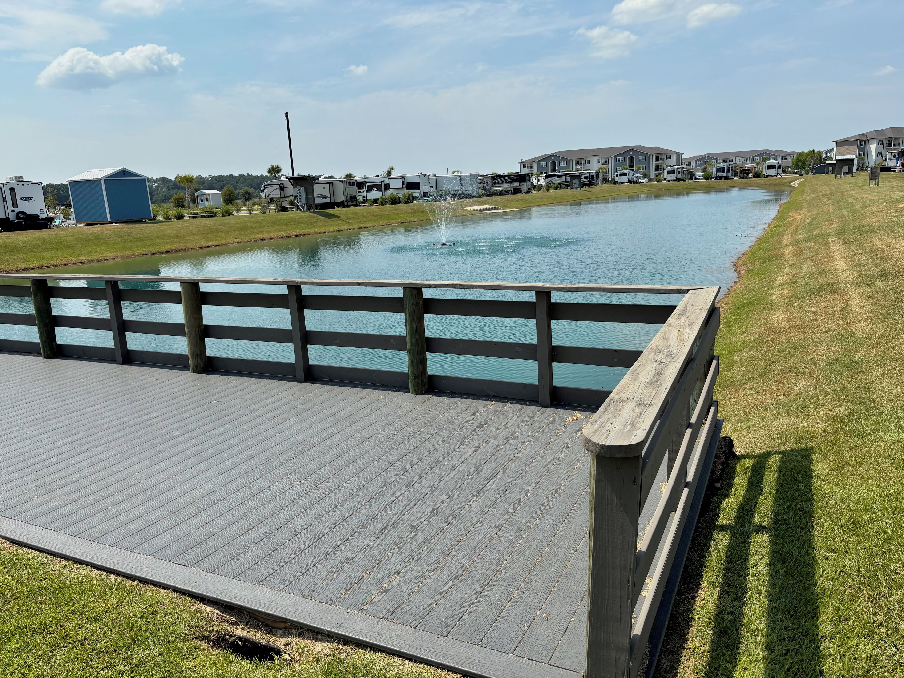A wooden deck with a railing overlooking a calm body of water, surrounded by grass and a row of houses.