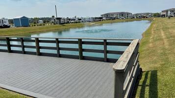 A wooden deck with a railing overlooking a calm body of water, surrounded by grass and a row of houses.