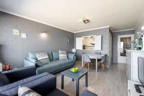 Ein modernes Wohnzimmer mit einem grauen Sofa, einem Essbereich mit Tisch und Stühlen sowie einer durch eine offene Tür einsehbarer Küche.