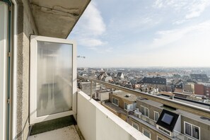 Ein Balkon mit Blick auf eine Stadtlandschaft, ein Fenster mit Jalousien und ein Metallgeländer.