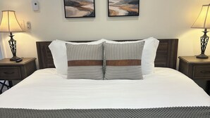 A neatly made bed with a white duvet, two pillows with striped patterns, and a grey textured throw. There are two bedside tables with lamps, and two framed artworks on the wall above the bed. - Cozy 3-bed mtn condo in village, walkable to all! (Slaty Fork)