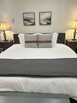 A neatly made bed with a white duvet, two pillows with striped patterns, and a grey textured throw. There are two bedside tables with lamps, and two framed artworks on the wall above the bed. - Cozy 3-bed mtn condo in village, walkable to all! (Slaty Fork)