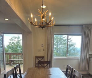 A dining area with a chandelier, a large table, and chairs. There are windows with curtains, and a view of trees outside. - Cozy 3-bed mtn condo in village, walkable to all! (Slaty Fork)