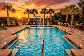 A swimming pool with a water feature, surrounded by palm trees and lounge chairs, with a sunset in the background. - New Domani Resort Escape | Minutes to Disney (Davenport)