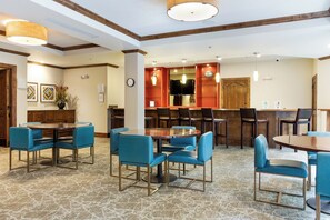 Eine Hotelhalle mit mehreren Sitzbereichen, darunter runde Tische und Stühle, einer Bar und einem Empfangstresen.