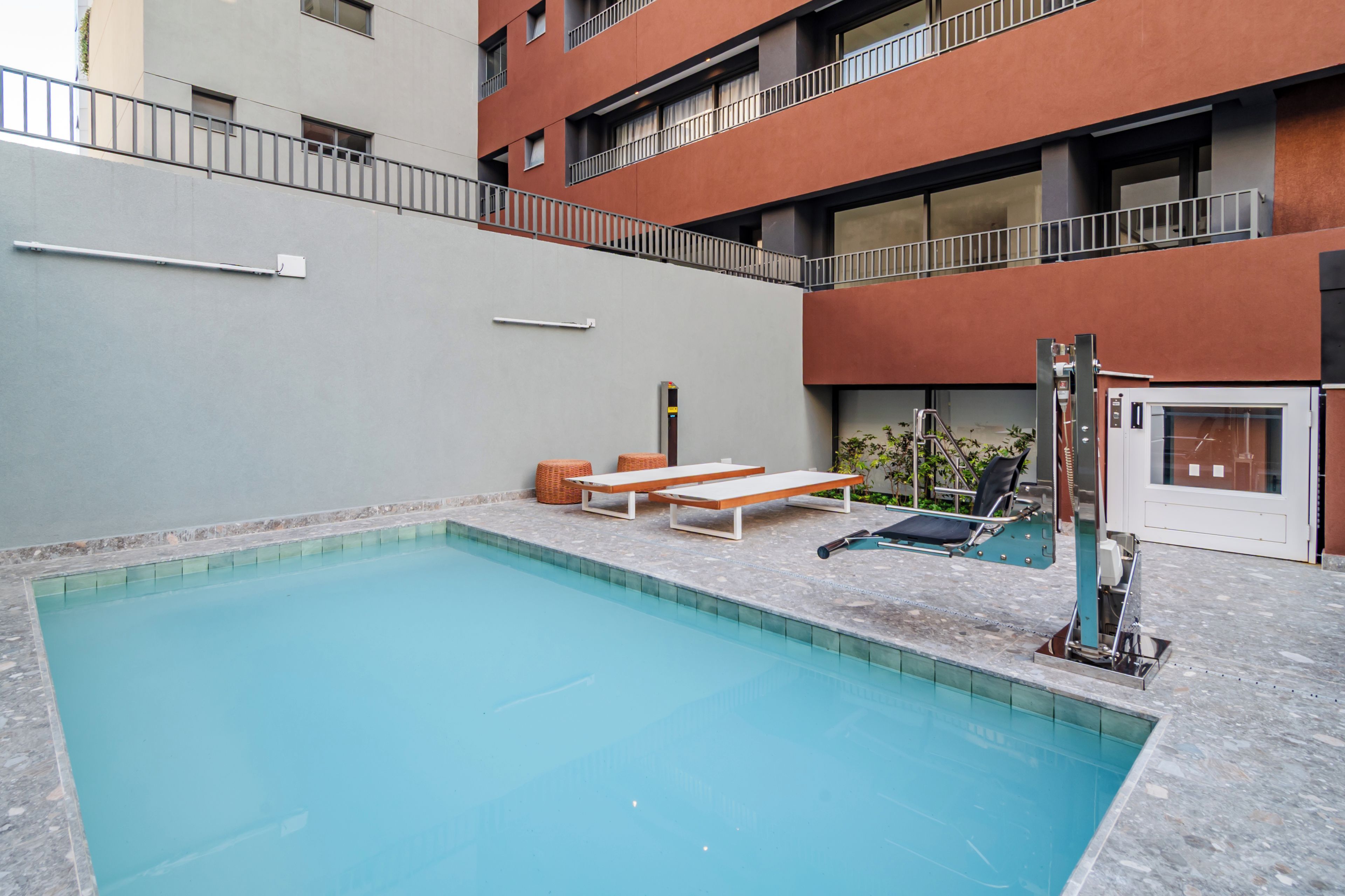 A swimming pool with a bench and lounge chairs, surrounded by a building with balconies.
