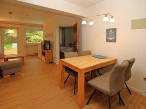 A room with a wooden dining table, two chairs, a bench, a television on a stand, and a door leading to another room. - Flurstrasse 10, Holiday Apartment 1 (Wyk auf Foehr)