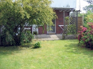 Un bâtiment en briques doté d’un porche couvert, d’un petit jardin avec de la pelouse et des arbustes, et d’un portail en métal.
