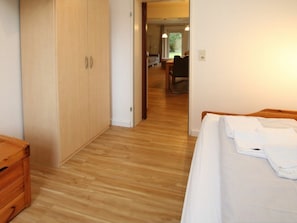 Une chambre avec un plancher en bois, un lit recouvert de draps blancs, une commode en bois et une porte donnant sur un espace de vie.