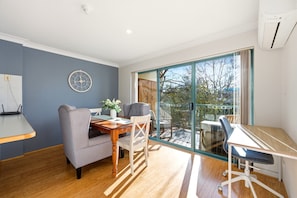 A modern dining area with a wooden table, grey upholstered chairs, and a large window offering a view of trees. - Stylish 2-bed Apartment in Kingston (Canberra)