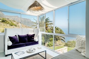 A modern living room with a sofa, a coffee table, and large windows offering a view of the sea and hills. - Camps Bay 6 Bed Ocean Views and Pool Villa Kai (Cape Town)
