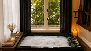 Um quarto de spa com uma grande banheira cheia de bolhas, uma janela com cortinas e velas ao lado.