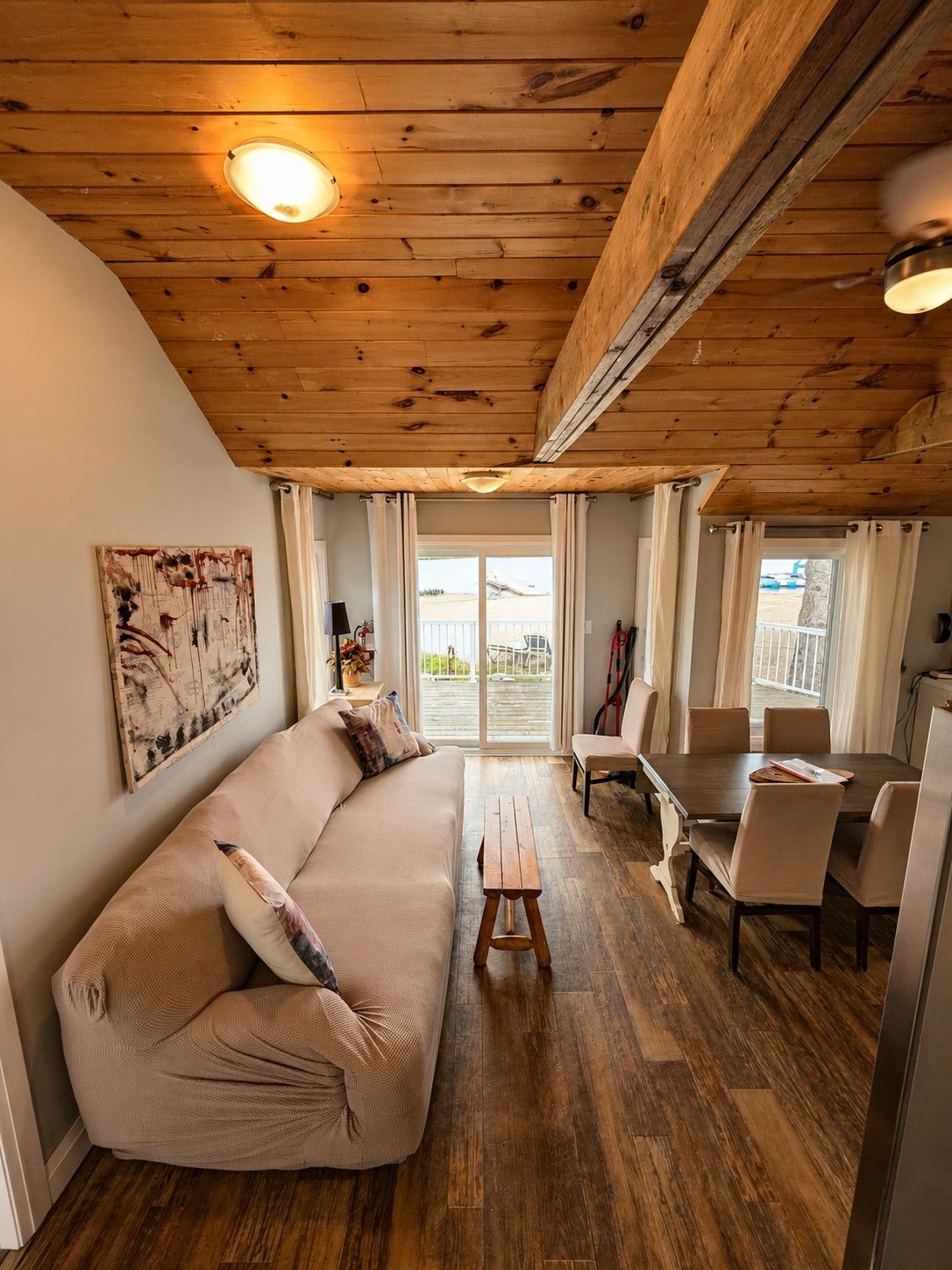 A cozy living area with a wooden ceiling, a sofa, a dining table with chairs, and a view through the glass doors.