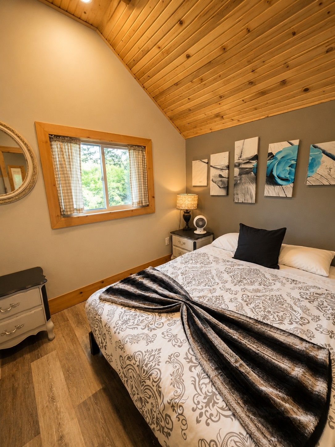 Un dormitorio con techo de madera, una cama con ropa de cama estampada, una mesita de noche con lámpara, un espejo y una obra de arte en la pared.