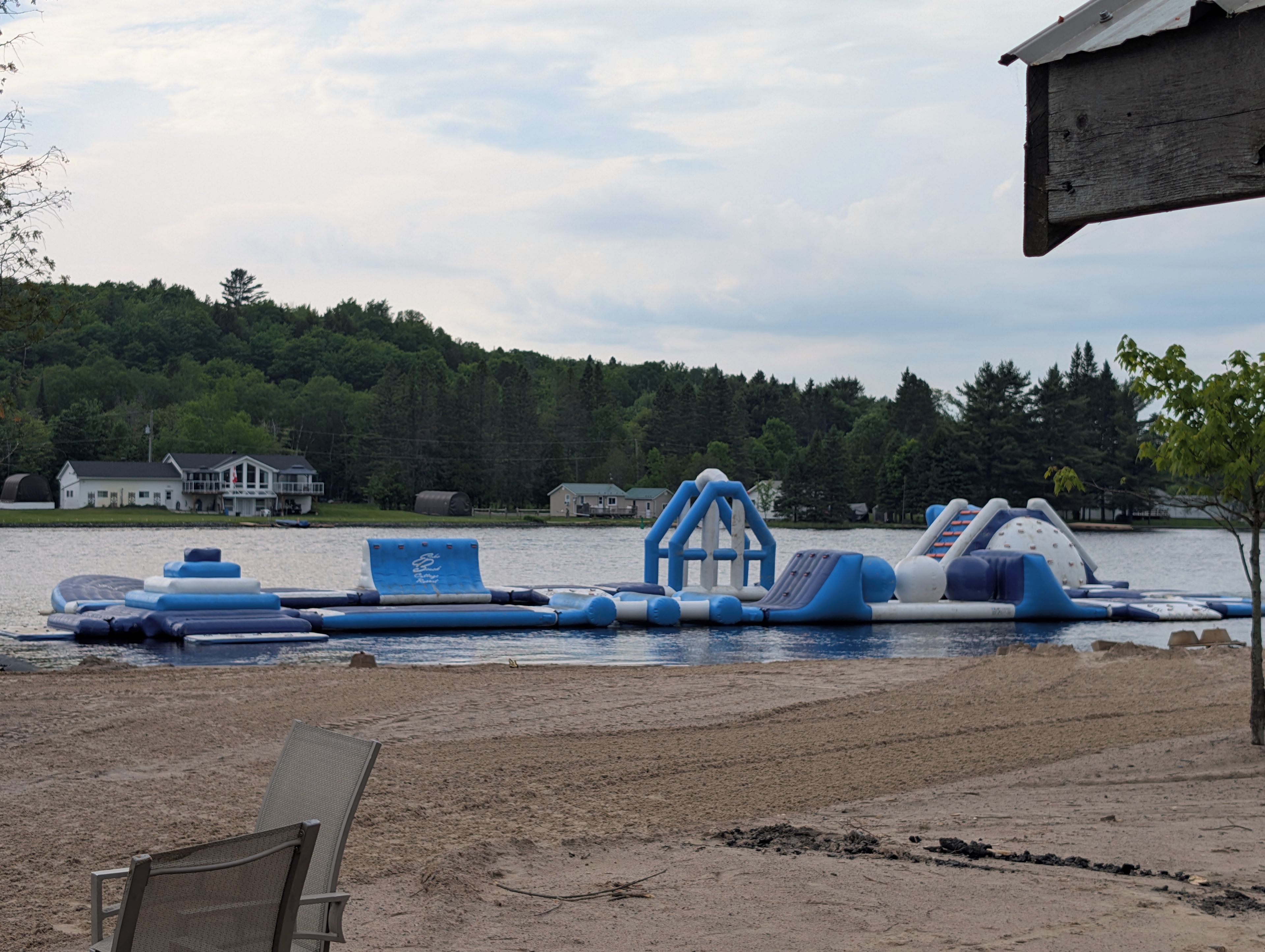 A lakeside setting with a sandy beach, inflatable water park equipment, and houses in the background.