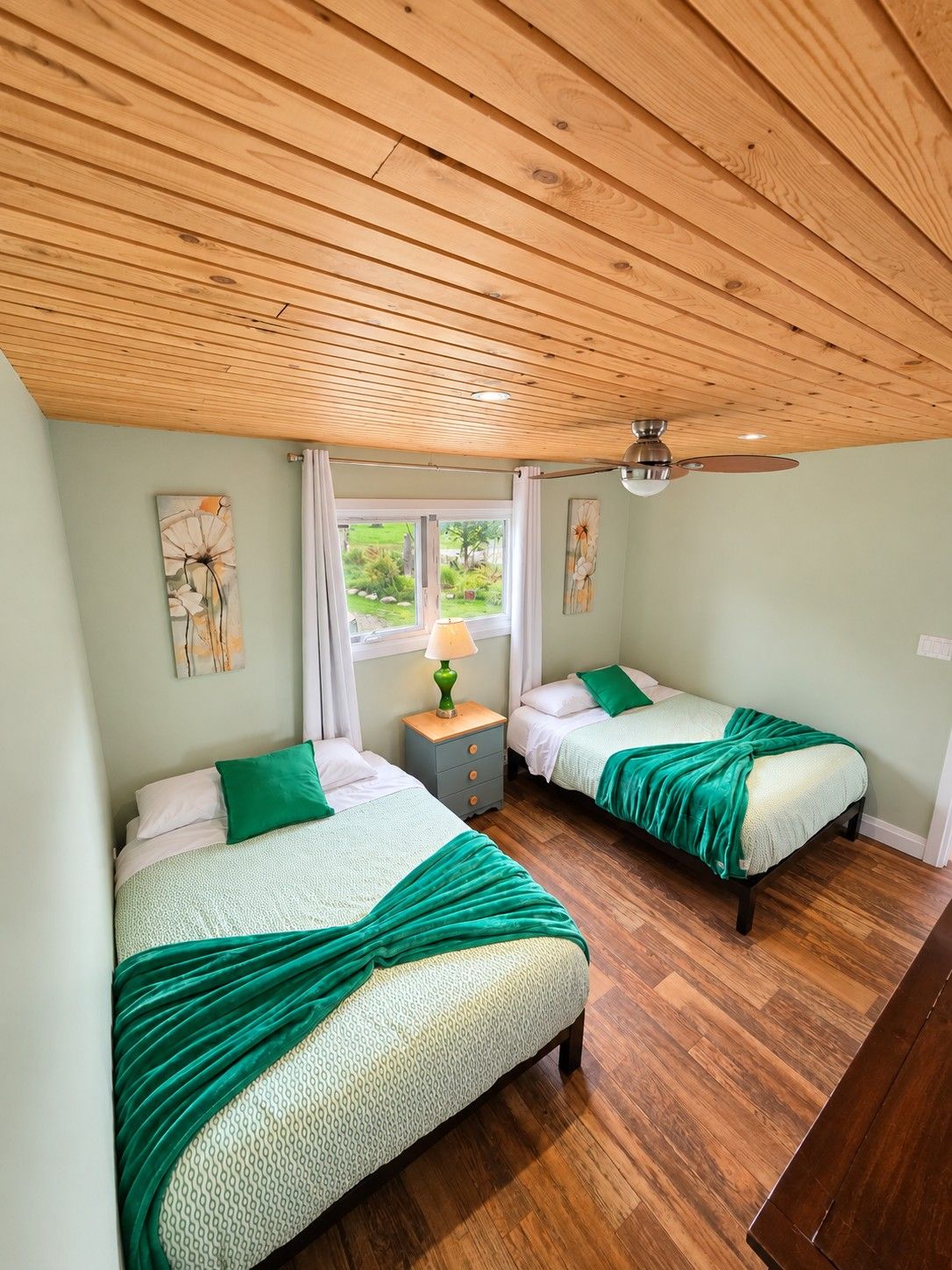 A room with two beds, wooden flooring, a ceiling fan, and artwork on the walls.