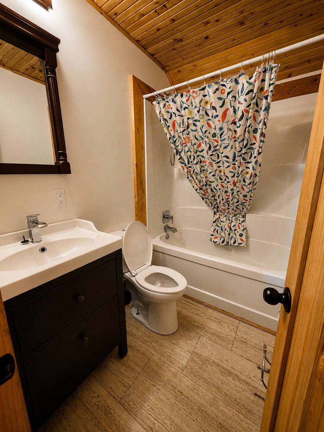 Un baño con techo de madera, un lavamanos blanco, un inodoro y una bañera con cortina de ducha estampada con motivos florales.