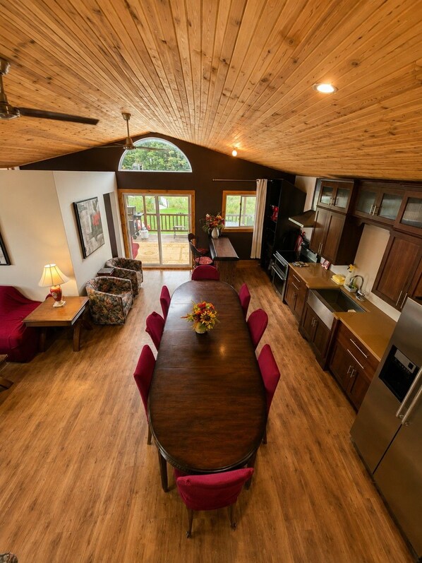 Un vaste espace à manger avec une table en bois et des chaises rouges, une cuisine équipée d’appareils électroménagers modernes, et une vue sur une terrasse en bois, visible à travers de grandes fenêtres.