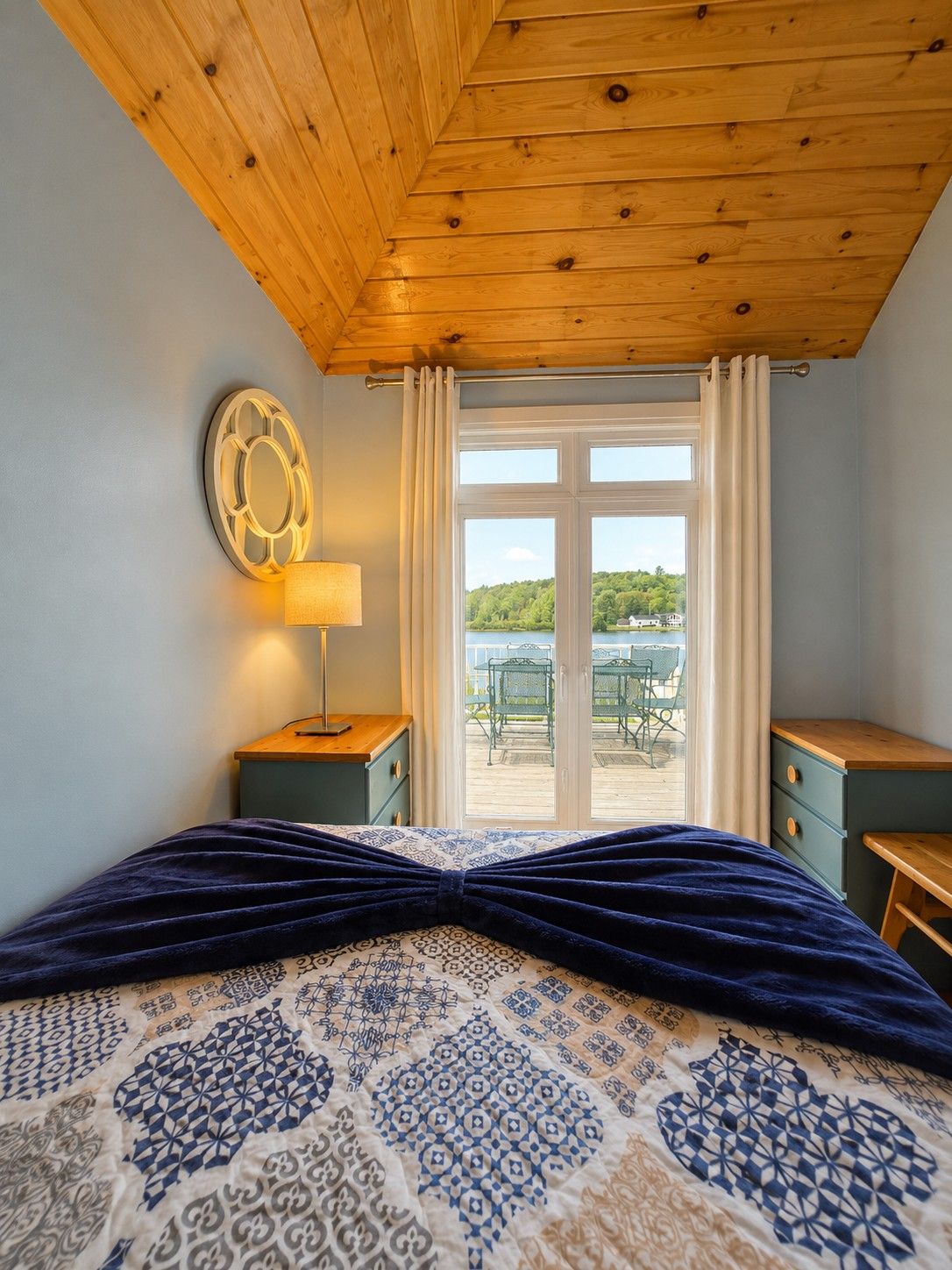 Un dormitorio con cama, mesita de noche, lámpara, ventana con cortinas y vista a un patio exterior.