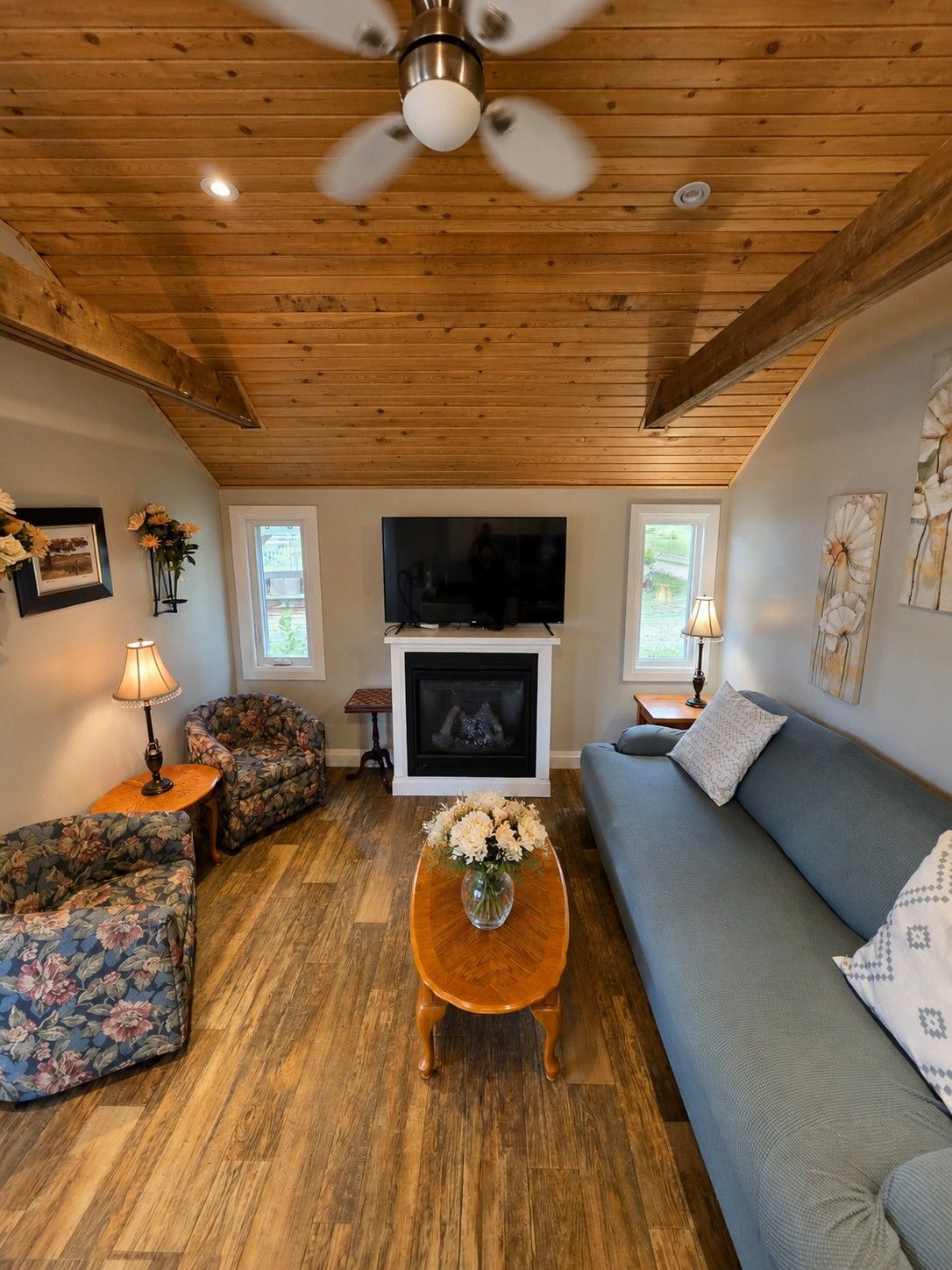 A living room with a wooden ceiling, a fireplace, a television, floral-patterned furniture, and a ceiling fan.