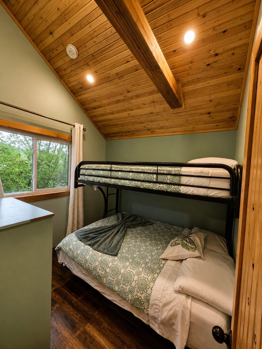 Un lit superposé avec un bureau et une fenêtre, dans une pièce dont le plafond est orné de poutres en bois.