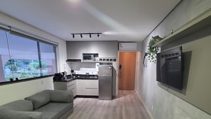 A modern kitchen with a refrigerator, microwave, and sink. There is a gray sofa and a small table with a remote control. - Estúdio Premium ao lado do Iguatemi (Lago Norte)