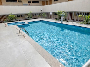 Una piscina rectangular con una escalera metálica en un lado, rodeada por un patio de baldosas y una valla metálica.